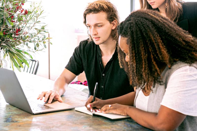 Two students working together at a table, one typing on a laptop and the other writing in a notebook